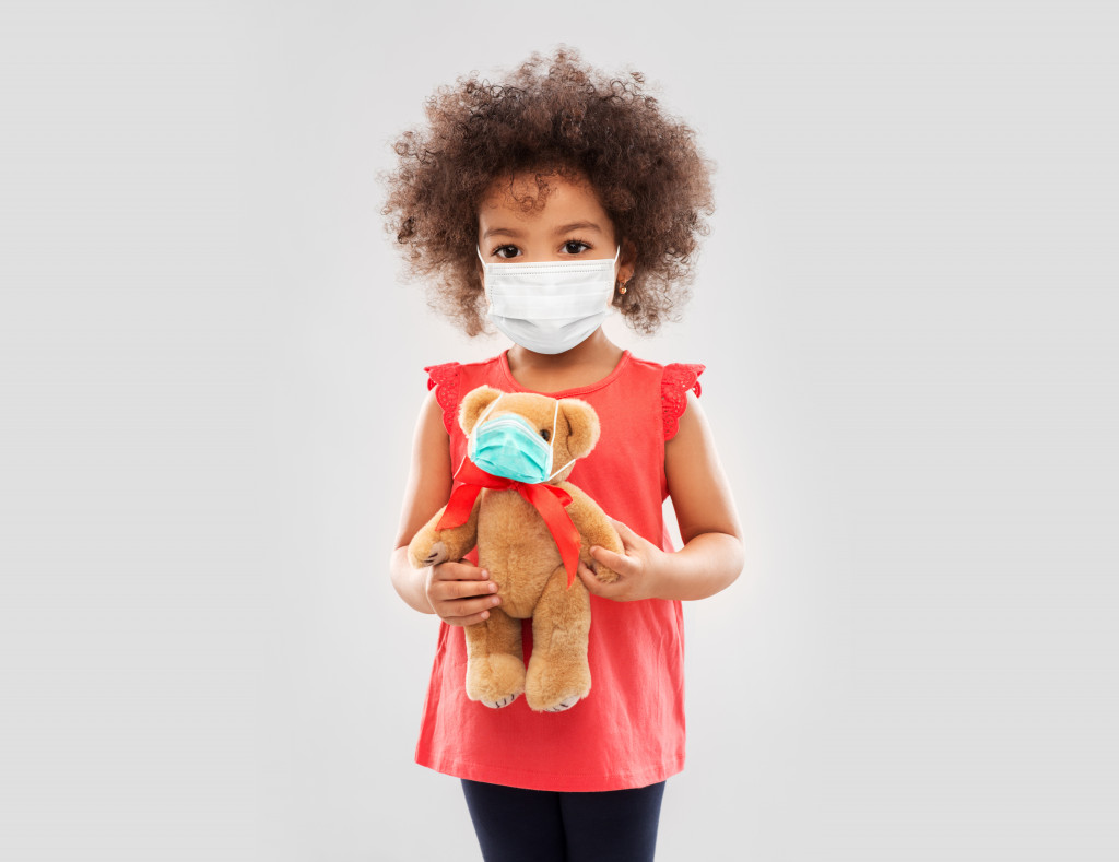 A young girl wearing a face mask while holding a teddy bear with a tiny mask