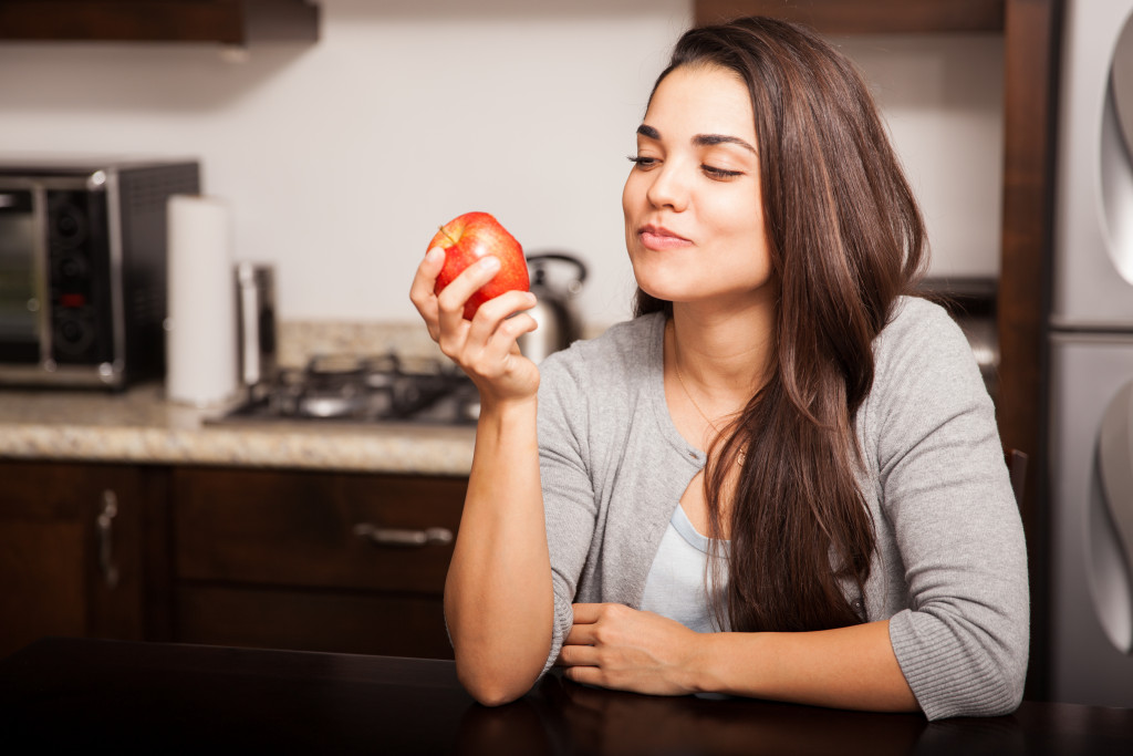 A woman in the kitchen holding and about to eat an apple
