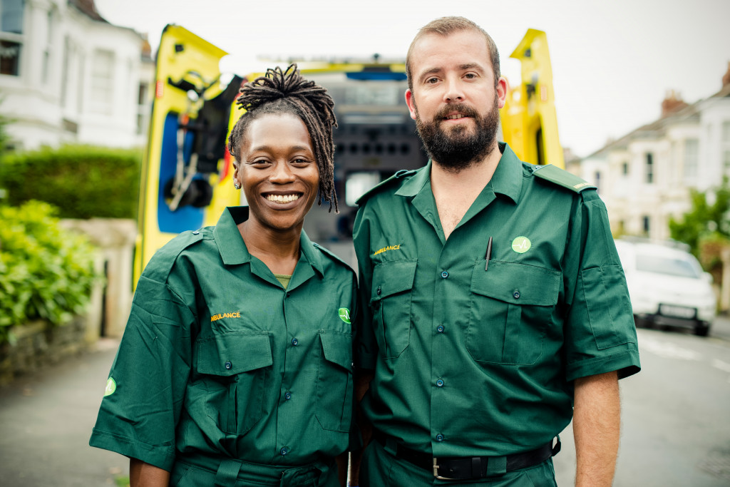 male and female paramedics wearing green uniforms in front of their truck smiling